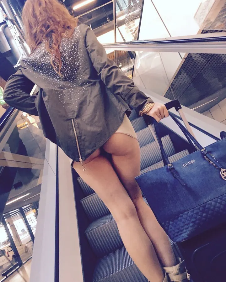 Femme en veste grise pailletée, montant un escalator avec une valise bleue