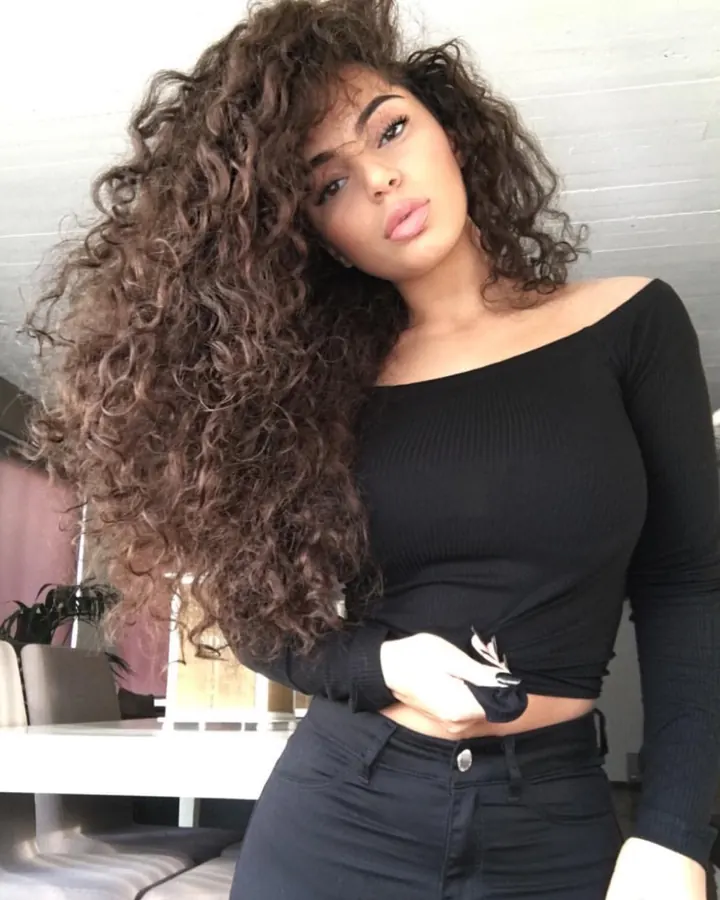 Femme aux cheveux bruns bouclés portant un haut noir ajusté, posant devant un mur blanc avec des plantes en arrière-plan.