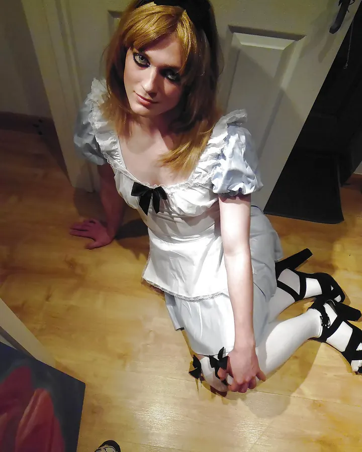 Femme blonde en robe blanche avec volants, accroupie sur un sol en bois, portant des collants blancs et des chaussures noires à brides.