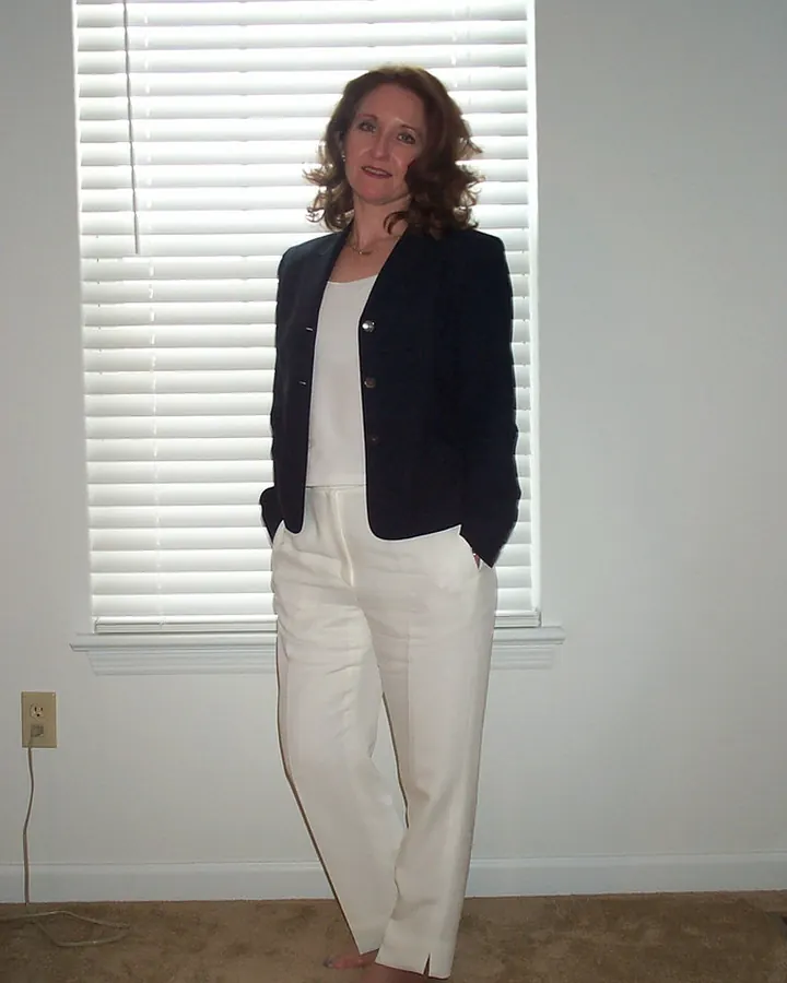 Femme aux cheveux châtains, en veste noire et pantalon blanc, debout devant une fenêtre avec des volets blancs, pieds nus.