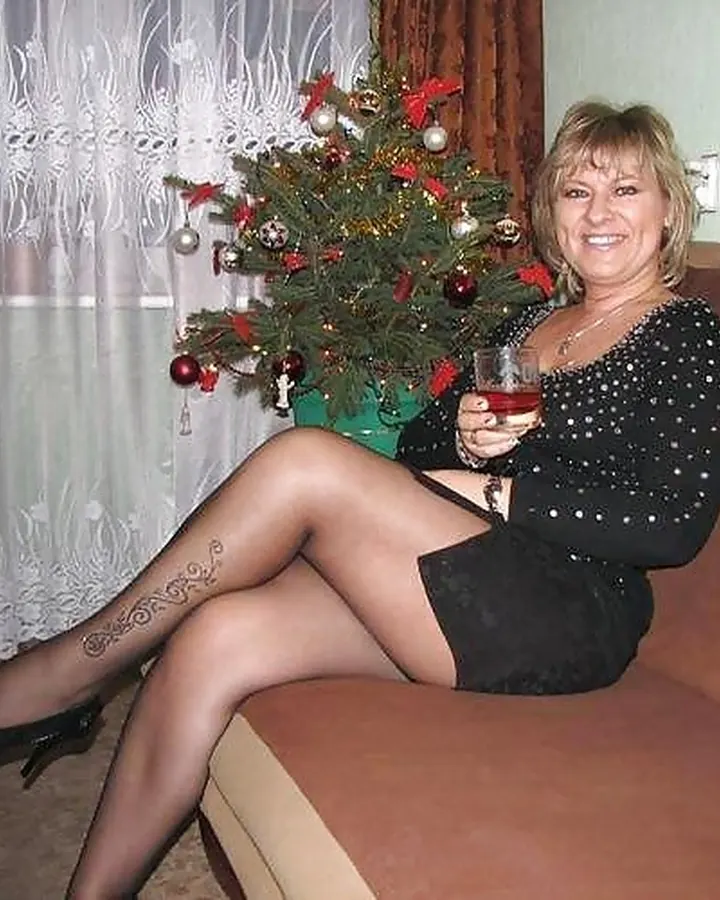 Femme aux cheveux blonds courts, portant une robe noire à pois, des collants foncés et des chaussures noires à talons, assise sur un canapé avec un verre à la main, près d’un sapin de Noël.