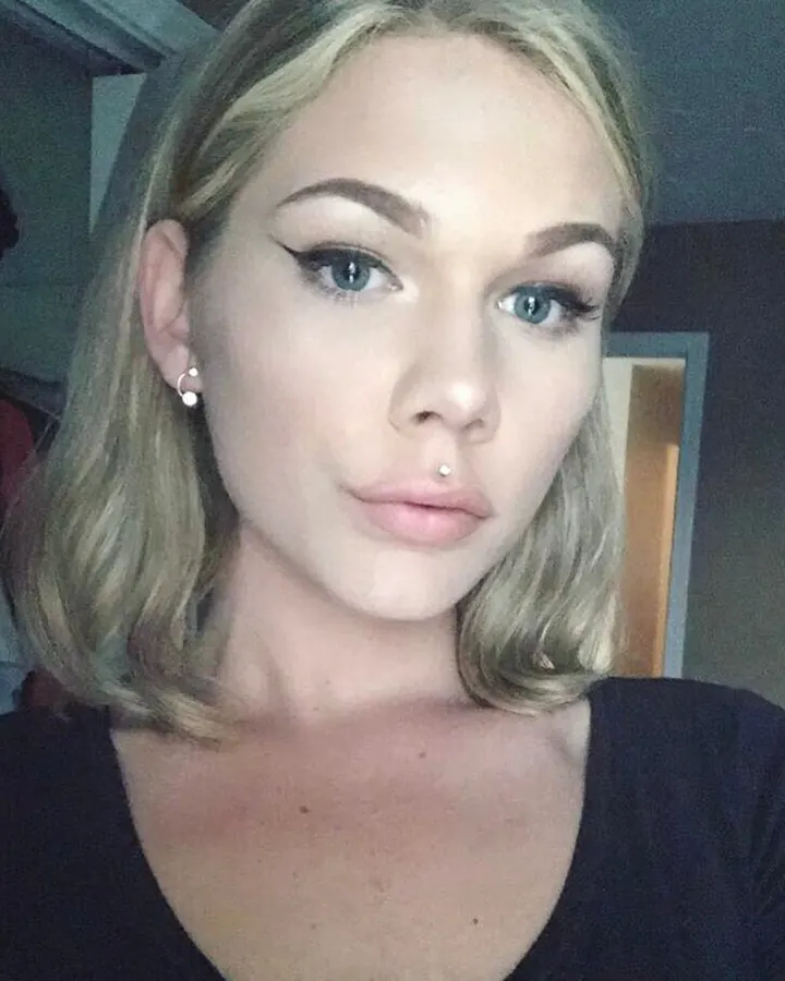 Femme aux cheveux blonds en haut noir, posant dans une chambre, regardant la caméra.