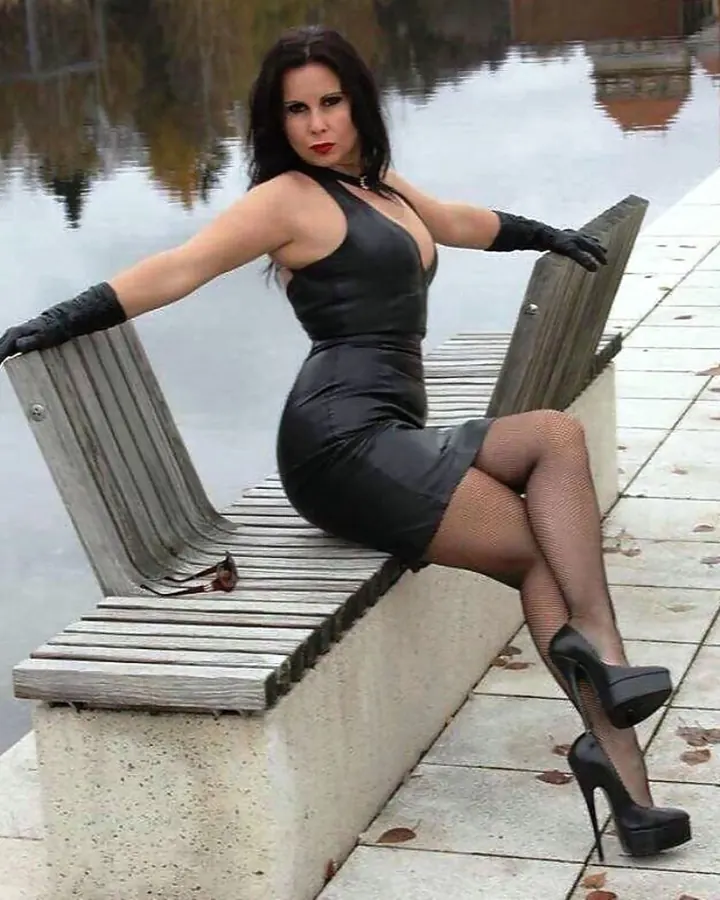 Femme aux cheveux foncés en robe noire, assise sur un banc en bois, près d'un lac, regardant l'appareil photo