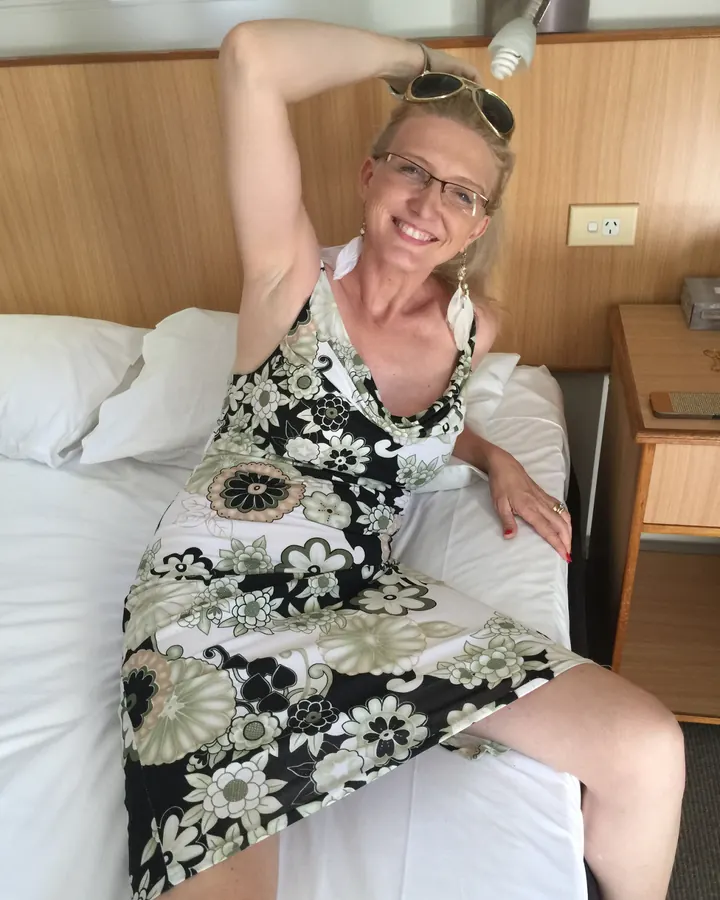 Femme blonde en robe à motifs floraux, allongée sur un lit, lunettes de soleil sur la tête, souriante.