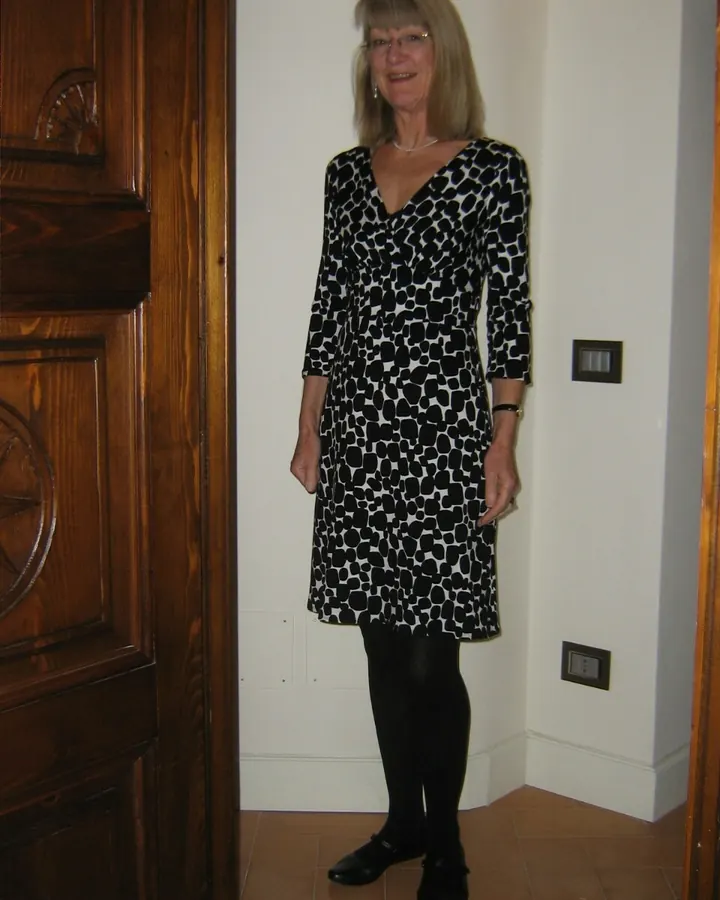 Femme blonde en robe noire et blanche à motifs géométriques, souriante, debout dans un couloir.