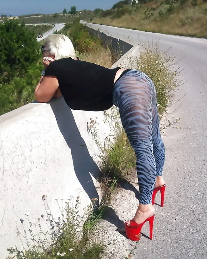 Femme blonde en tenue de cuir noir, debout près d'une voiture argentée, portant une jupe en jean courte et des escarpins noirs.