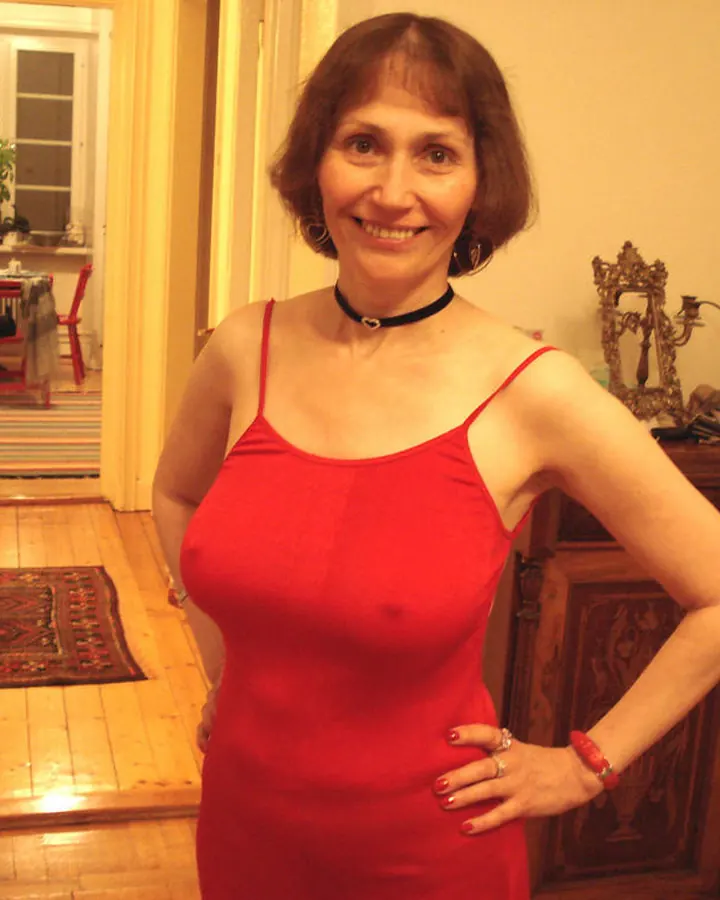 Femme aux cheveux châtains, en robe rouge, posant dans un salon avec des bougies rouges en arrière-plan.