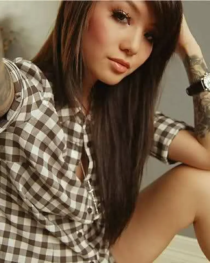 Femme aux cheveux bruns longs, tatouages aux bras, chemise à carreaux, pose assise sur un lit.