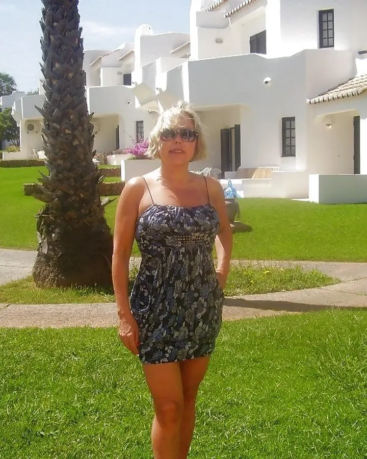 Femme aux cheveux blonds portant un haut noir, se tenant de face, regardant vers l'appareil, avec des lunettes noires et un collier doré en forme de lettre A.