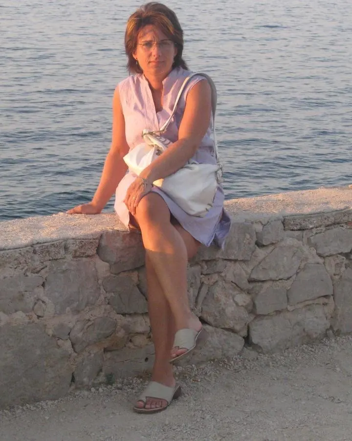 Femme aux cheveux bruns courts portant une robe lilas, assise sur un mur de pierre près de l'eau, tenant un sac blanc, pieds nus dans des sandales blanches.
