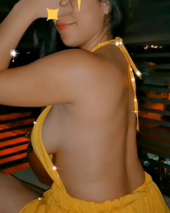 Femme aux cheveux bruns foncés portant un bikini rose pailleté, assise sur un lit avec une couverture colorée, les jambes écartées, les mains posées sur les cuisses.
