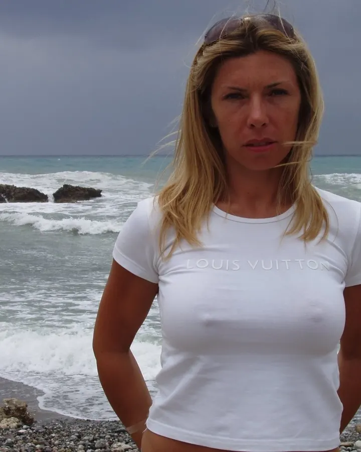 Femme aux cheveux blonds portant un haut noir, se tenant de face, regardant vers l'appareil, avec des lunettes noires et un collier doré en forme de lettre A.