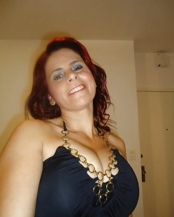 Femme aux cheveux roux portant une robe noire avec un collier en chaînes dorées, souriant à l'appareil photo