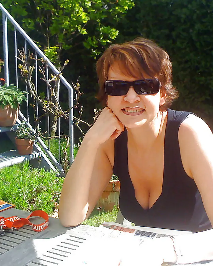 Femme en haut noir, assise à une table dehors, portant des lunettes de soleil, souriant à l'appareil photo