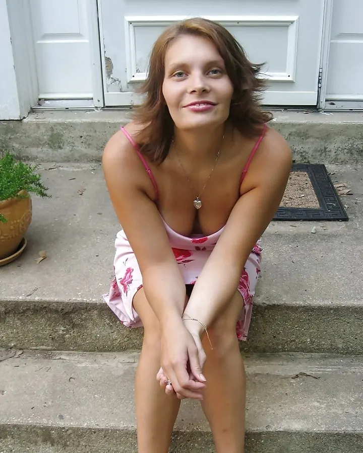 Femme brune assise sur des marches en béton devant une porte blanche, portant une robe rose à fleurs, souriant à la caméra.
