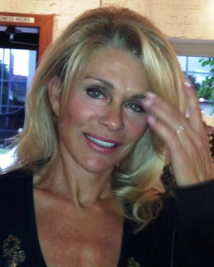 Femme blonde en vêtements sombres, souriant légèrement, main sur le front.