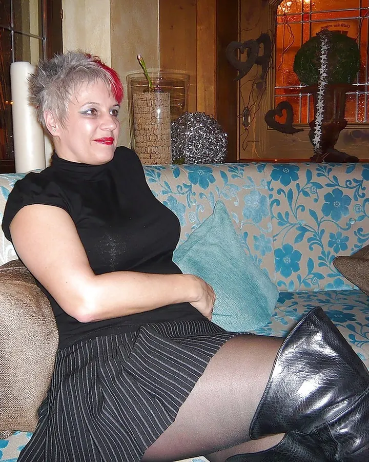Femme aux cheveux gris avec des mèches rouges, vêtue d'une robe noire et d'une jupe rayée, assise sur un canapé bleu et beige décoré de coussins.