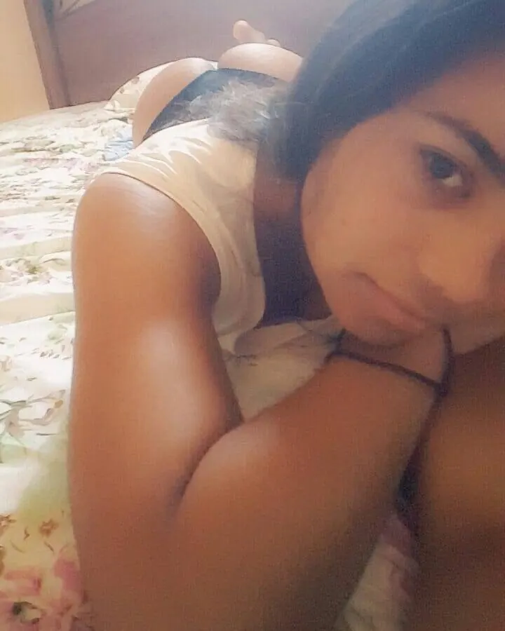 Femme allongée sur un lit, vêtue d’un haut blanc, regardant l’appareil photo avec un air neutre.