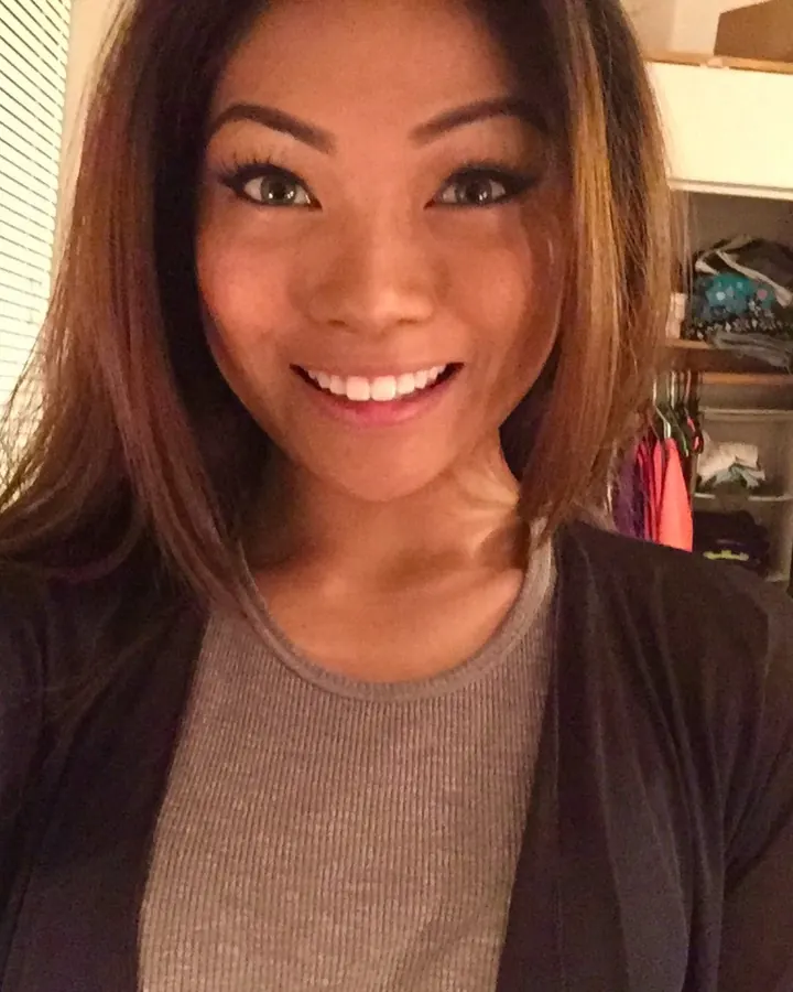Jeune femme aux cheveux bruns, souriante, en tenue décontractée, prenant un selfie dans une chambre.