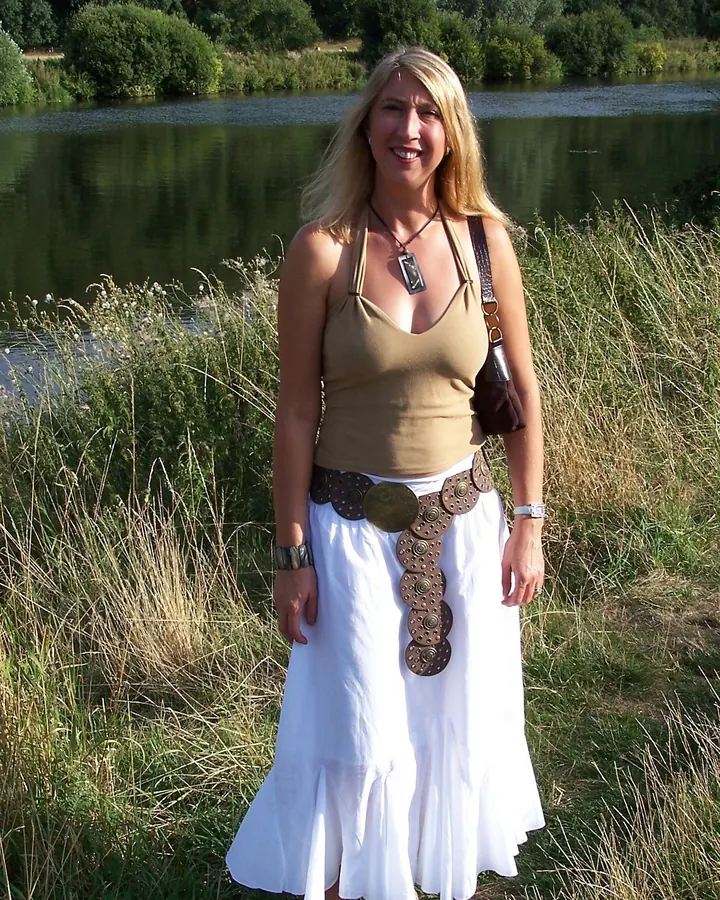 Femme blonde en tenue bohème, souriante, debout près d’un lac entouré de verdure.
