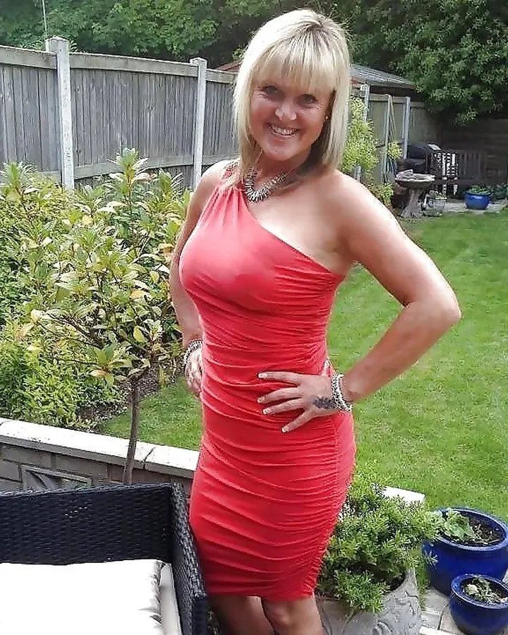 Femme blonde en robe rouge, souriante, posant dans un jardin avec des plantes et une terrasse.