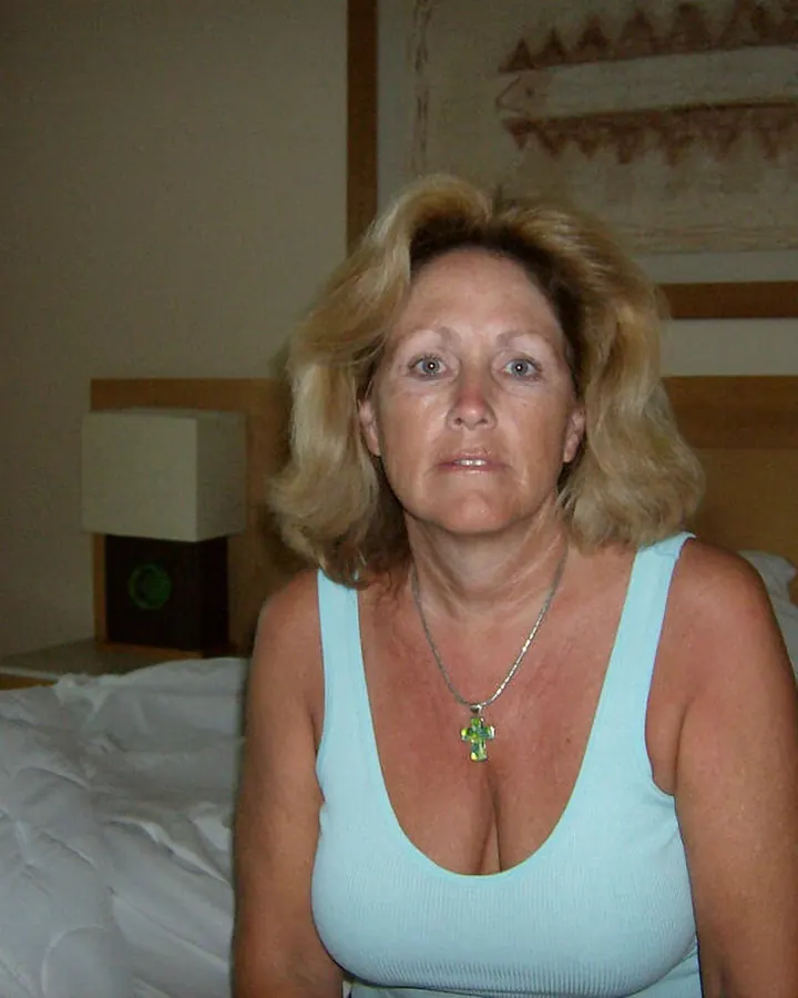 Femme aux cheveux bruns, en tenue blanche, assise sur une chaise métallique devant une fenêtre, souriante.