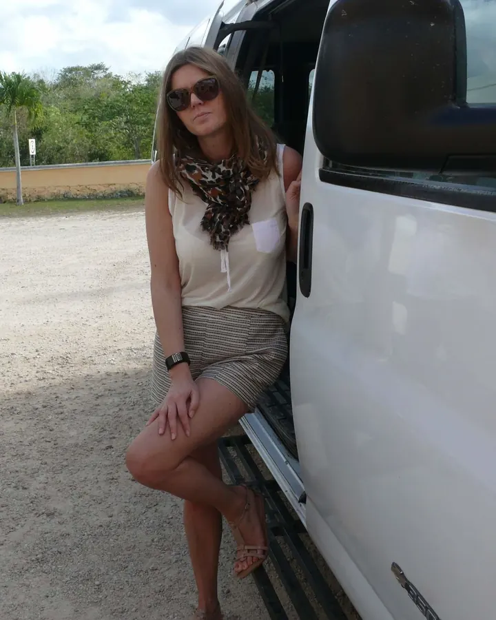Femme aux cheveux bruns en chemisier noir, jupe courte, posant près d'une voiture blanche