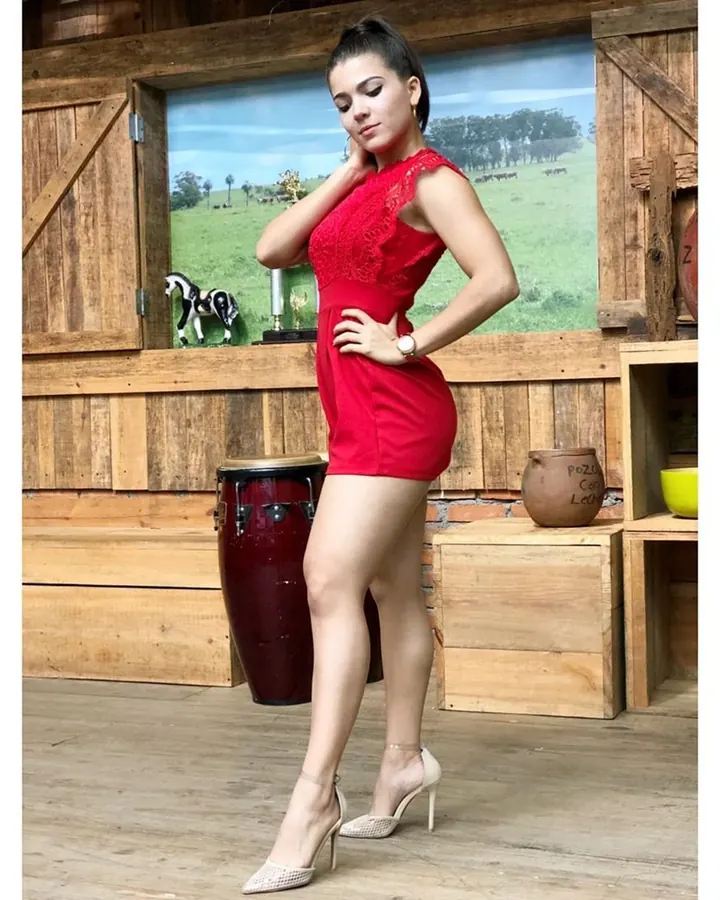 Jeune femme européenne aux cheveux bruns-noirs, portant une robe rouge ajustée, posant dans un décor en bois rustique avec un tambour en arrière-plan.