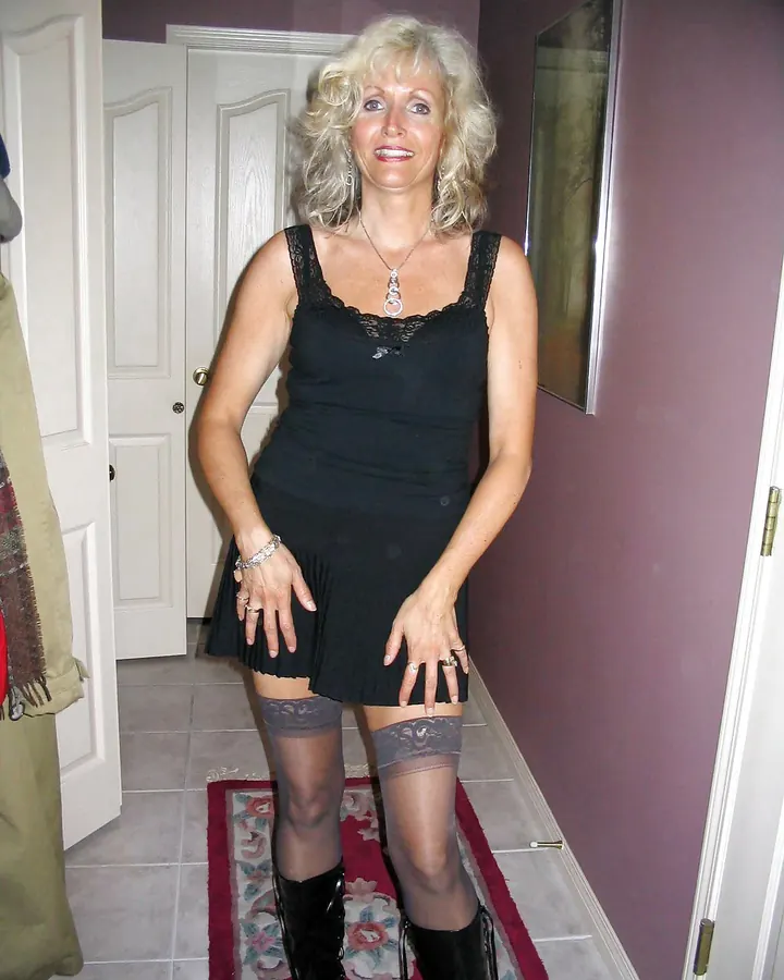 Femme européenne aux cheveux platine, portant une robe noire courte avec des bas résille et des bottes noires, debout dans un couloir intérieur.