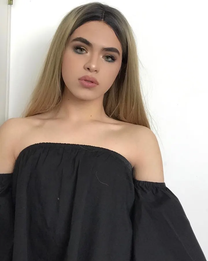 Jeune femme latino aux cheveux platine, portant un haut noir décolleté, posant devant un mur blanc, regardant vers le côté.