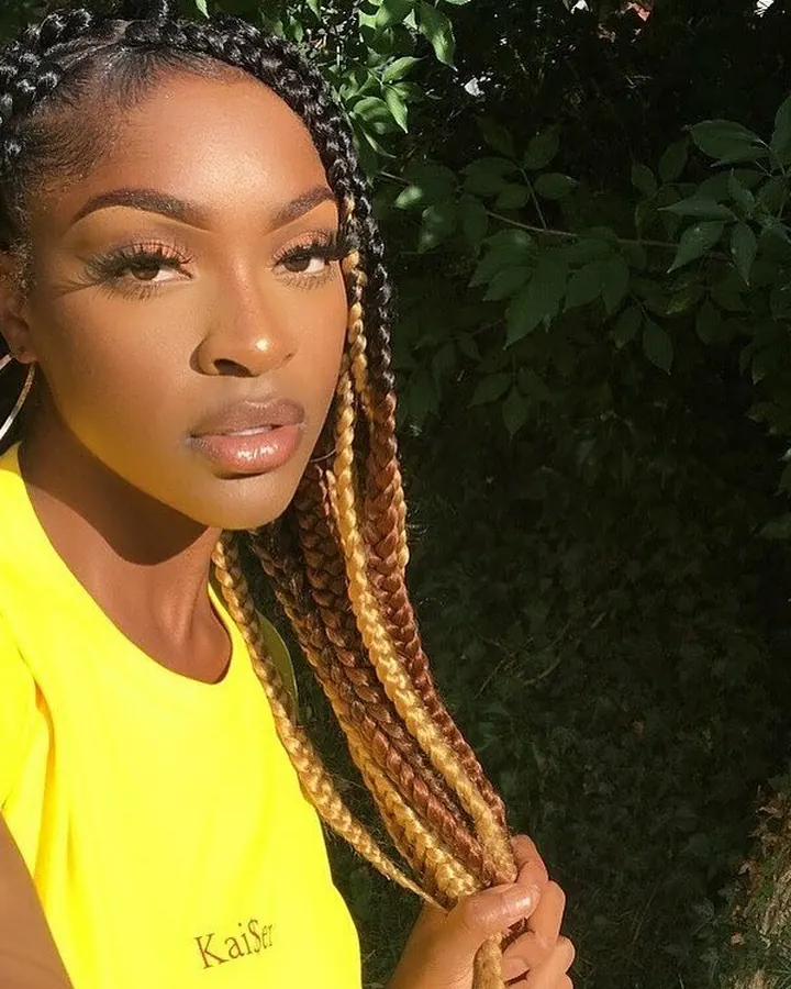 Femme africaine aux cheveux tressés, portant un t-shirt jaune vif, en extérieur devant des feuilles vertes.