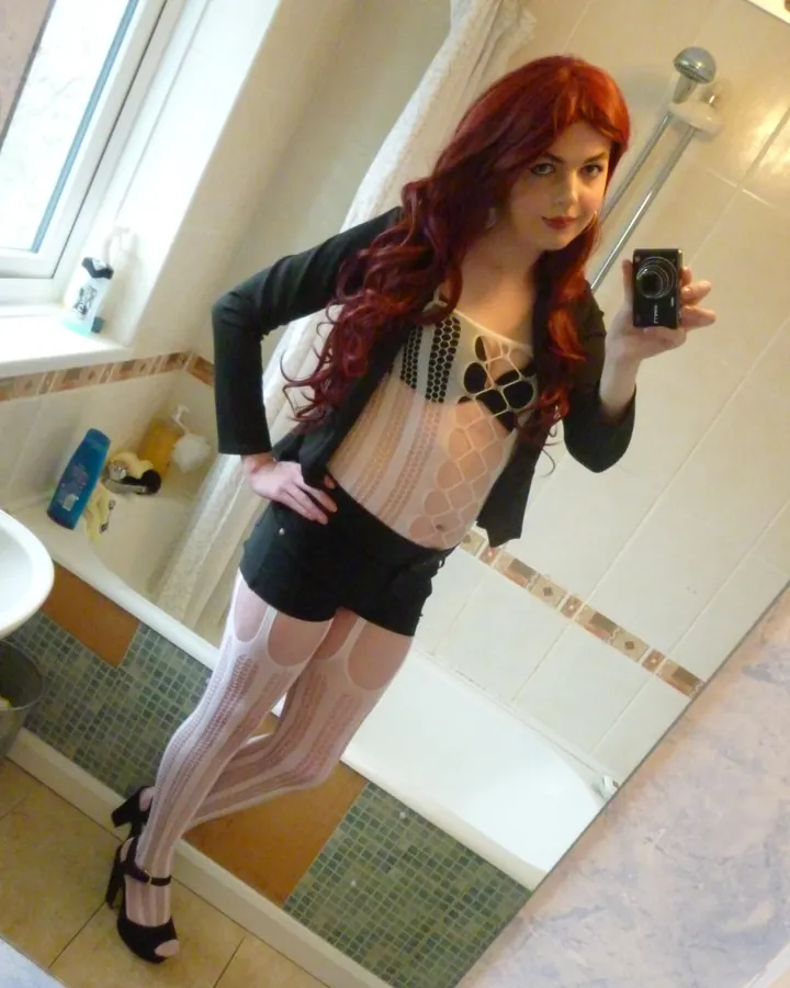 Femme aux cheveux roux, en lingerie blanche, debout devant une fenêtre, regard direct.