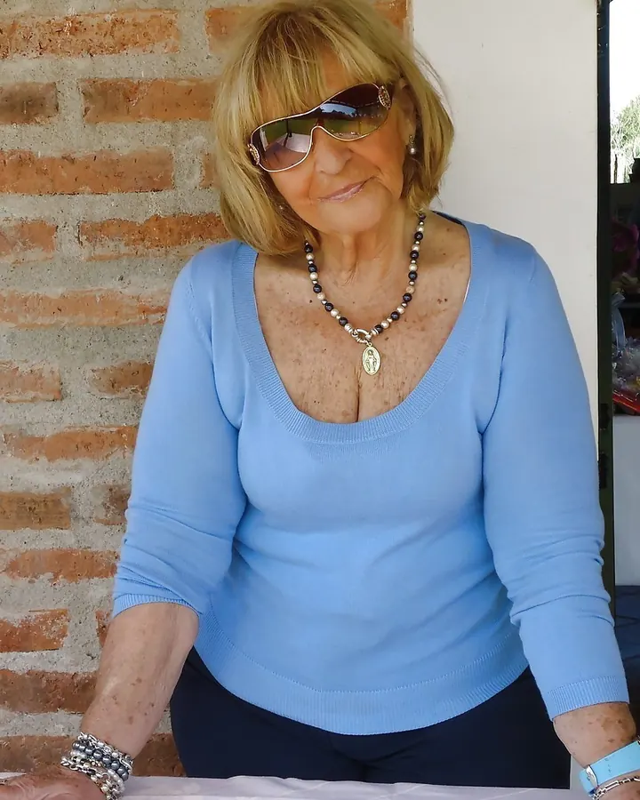 Femme blonde en pull bleu, assise contre un mur de briques, portant des lunettes de soleil, souriant légèrement.