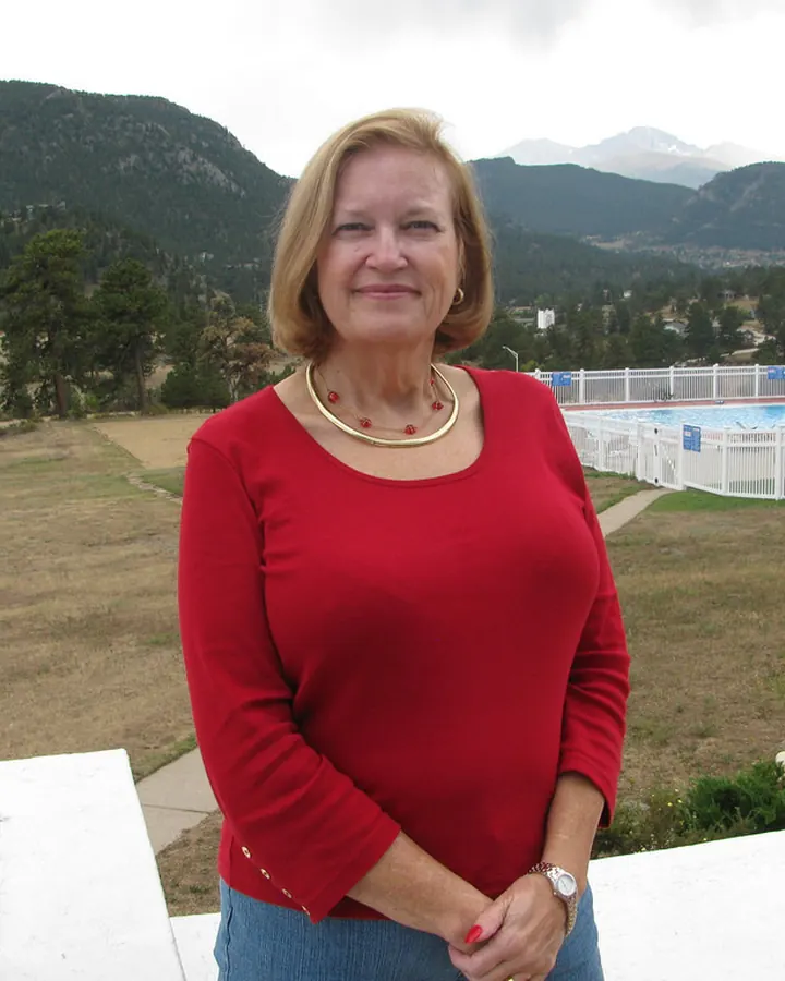 Femme aux cheveux blonds portant un pull rouge, souriant, debout sur une terrasse avec une vue sur des montagnes et une piscine.