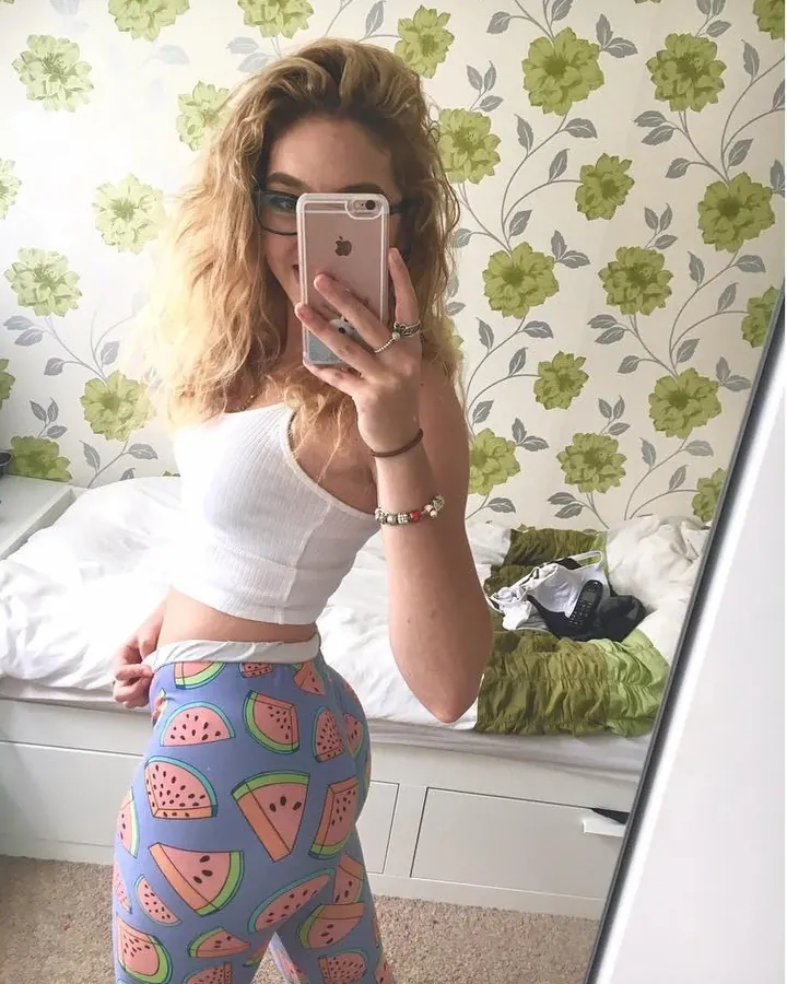 Femme aux cheveux bruns, souriante, en haut bleu clair, prenant un selfie dans un couloir.