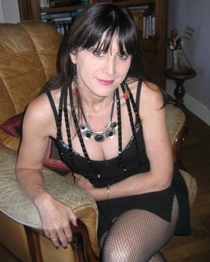Femme aux cheveux noirs mi-longs, regard direct, portant un haut noir avec collier métallique, assise sur un canapé en cuir.
