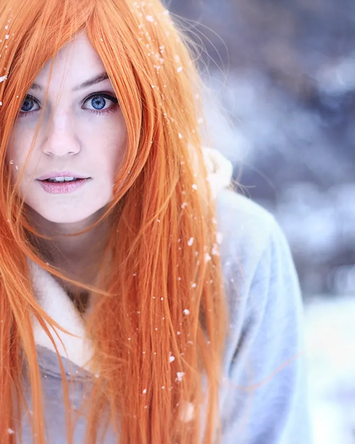 Femme aux cheveux roux, en pull gris clair, posant dans la neige, regardant directement l'appareil, des flocons sur les cheveux.