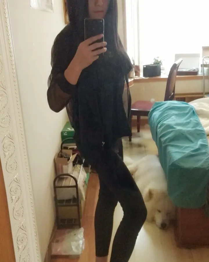 Jeune femme aux cheveux noirs longs, vêtue d'un haut noir transparent et de leggings noirs, prenant un selfie devant un miroir dans une chambre.