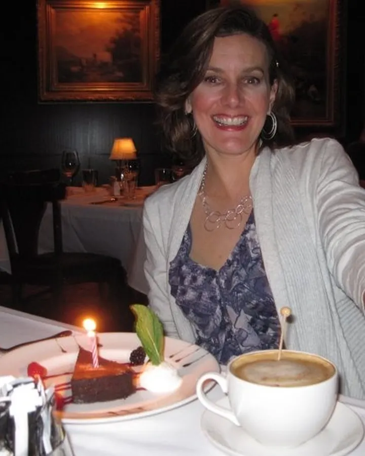 Femme aux cheveux blonds, souriante, assise à une table avec un gâteau d'anniversaire et une tasse de café.