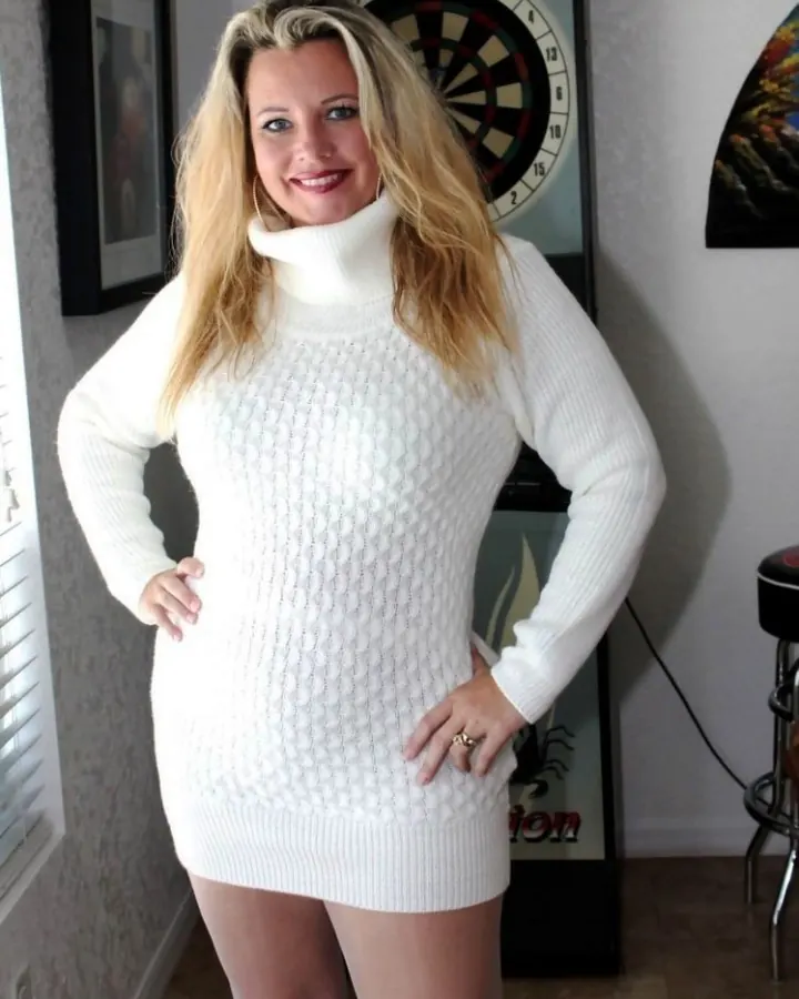 Femme blonde, vêtue d'un pull blanc col roulé, posant devant un jeu de fléchettes.