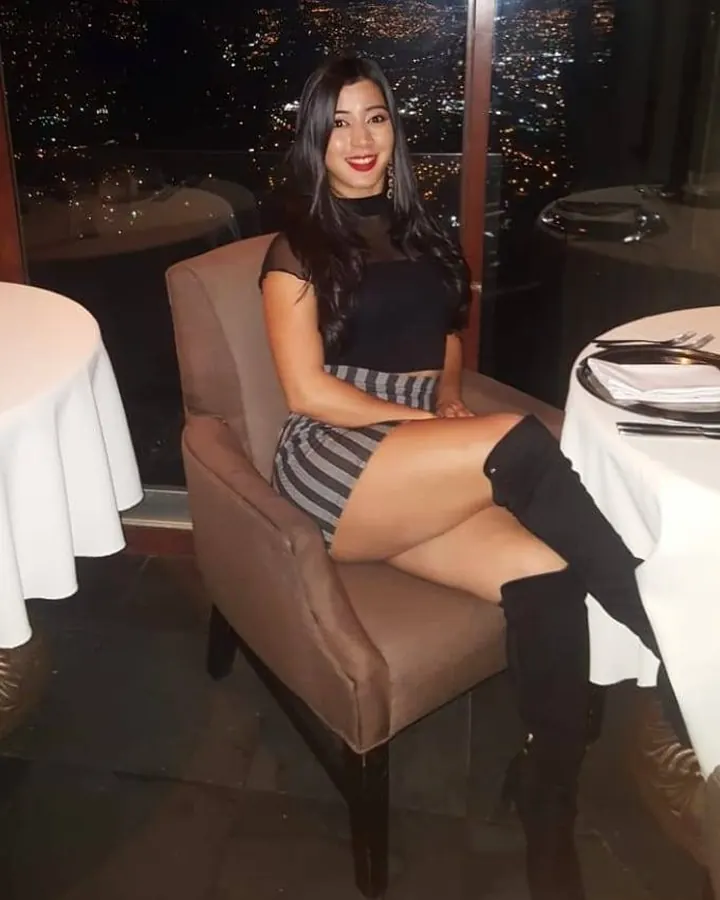 Femme asiatique souriante, assise dans un restaurant avec vue sur la ville la nuit, portant une robe noire et des bottes hautes.