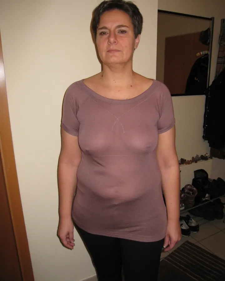 Femme aux cheveux bruns en robe rose, debout près d’une porte blanche, les mains sur les hanches.