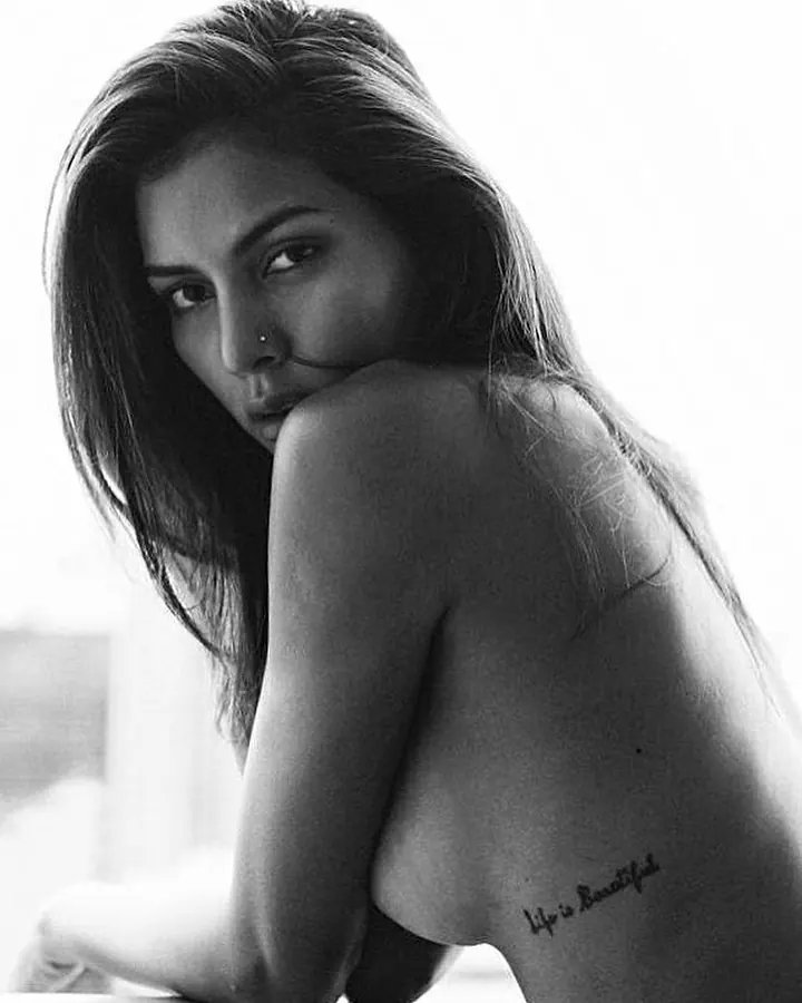 Photo en noir et blanc d'une femme aux cheveux foncés, nue, regardant l'objectif, avec un tatouage 'Life is Beautiful' sur le dos.
