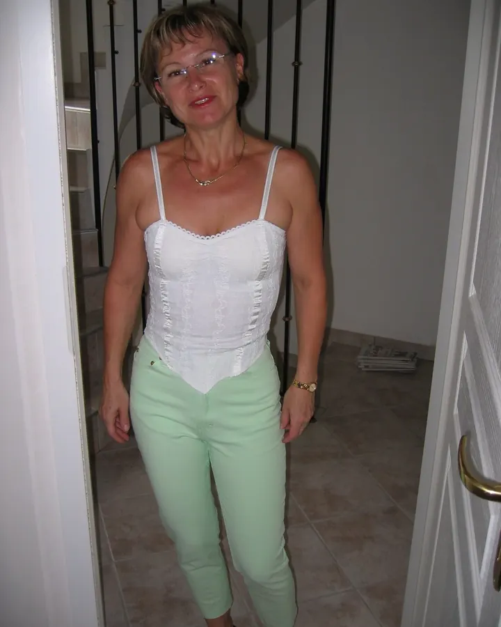 Femme aux cheveux courts blonds, portant un haut blanc et un pantalon vert clair, debout dans un couloir.