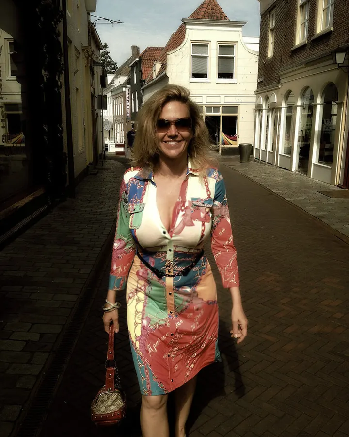 Femme souriante en robe colorée, lunettes de soleil, rue pavée, sac à main