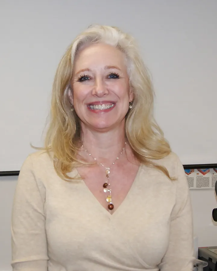 Femme aux cheveux blonds et yeux bleus, souriante, portant une tenue noire et bleue, assise à une table en extérieur.