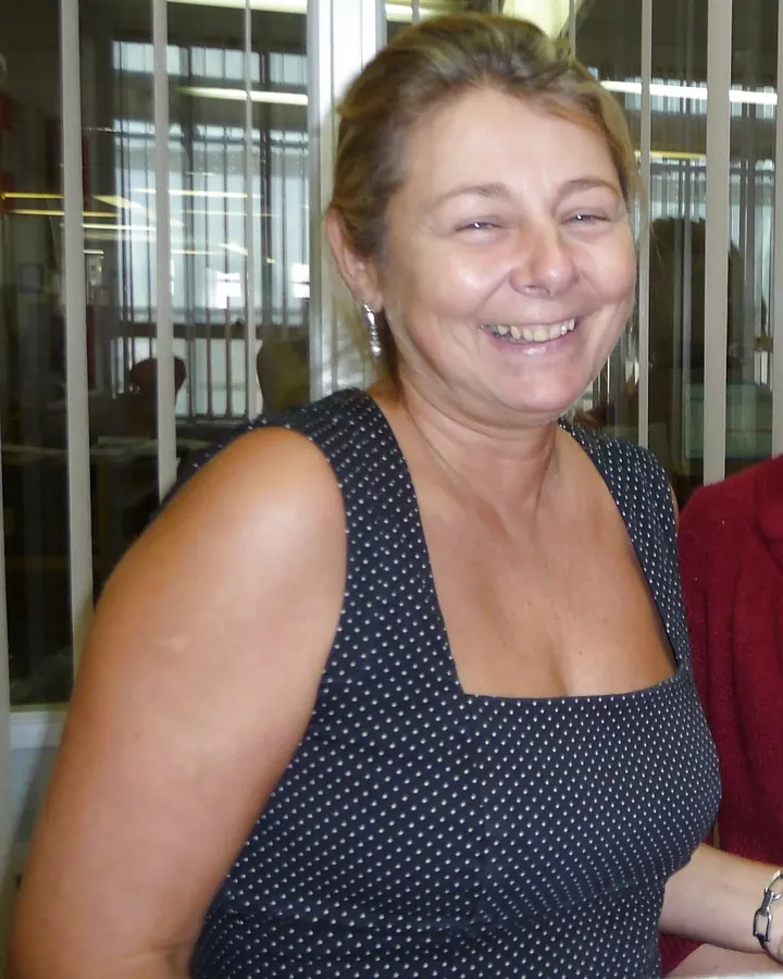 Femme aux cheveux blonds et yeux marrons, souriante, portant un haut blanc, dans une cuisine.