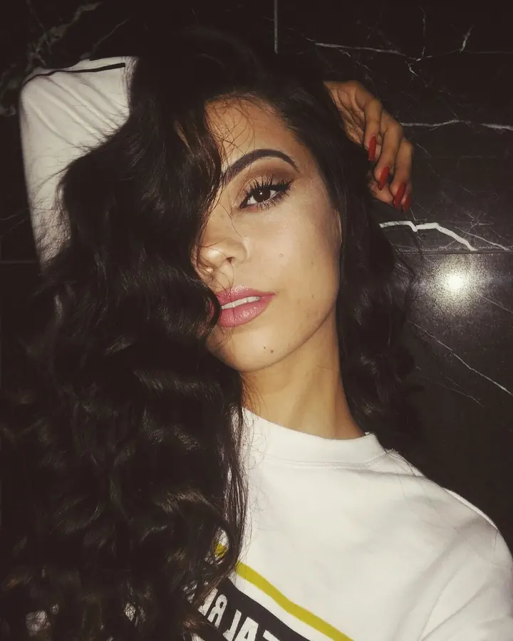 Femme aux cheveux bruns noirs en haut blanc, allongée sur un lit, regardant l'objectif avec un sourire.