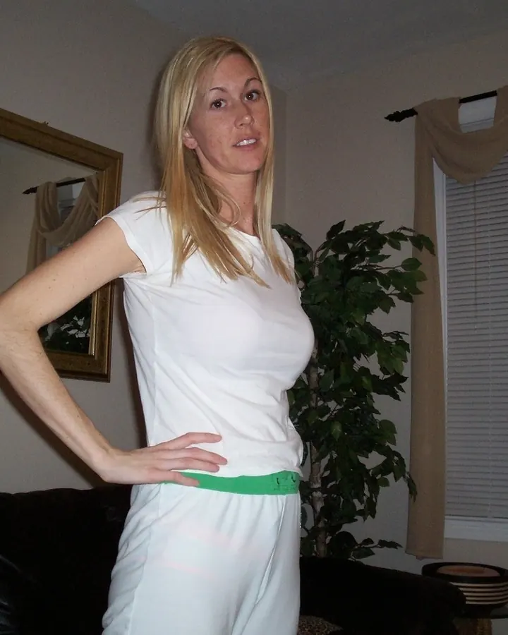 Femme blonde en tenue blanche, debout dans un salon, main sur la hanche, regardant vers le haut