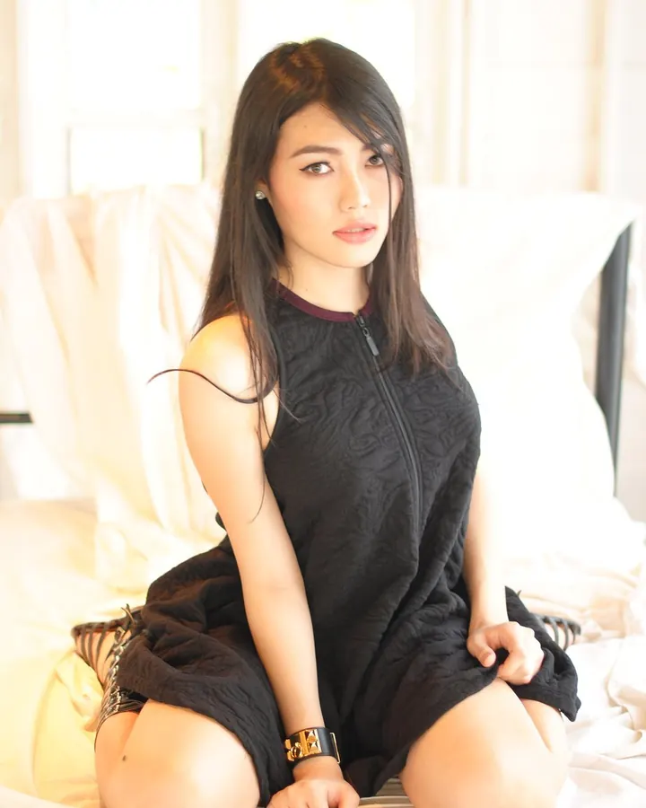 Jeune femme asiatique en robe noire, assise sur un lit blanc, regardant vers la caméra, bracelet doré au poignet